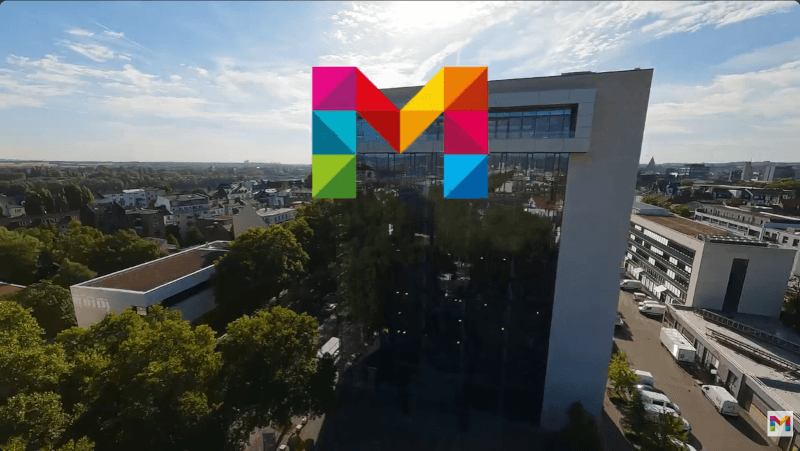 Luftaufnahme eines modernen Bürogebäudes mit Glasfassade inmitten einer Stadtlandschaft. Im Vordergrund ist das bunte M zu sehen.